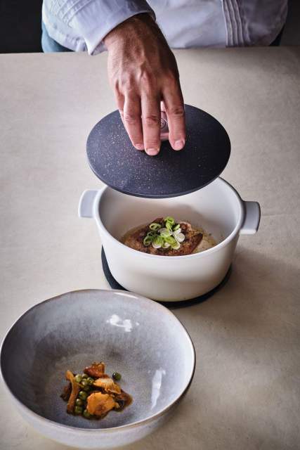 Main soulevant le couvercle d'une cocotte avec bouillon et garniture en assiette creuse – La Table des Échevins Restaurant Gastronomique